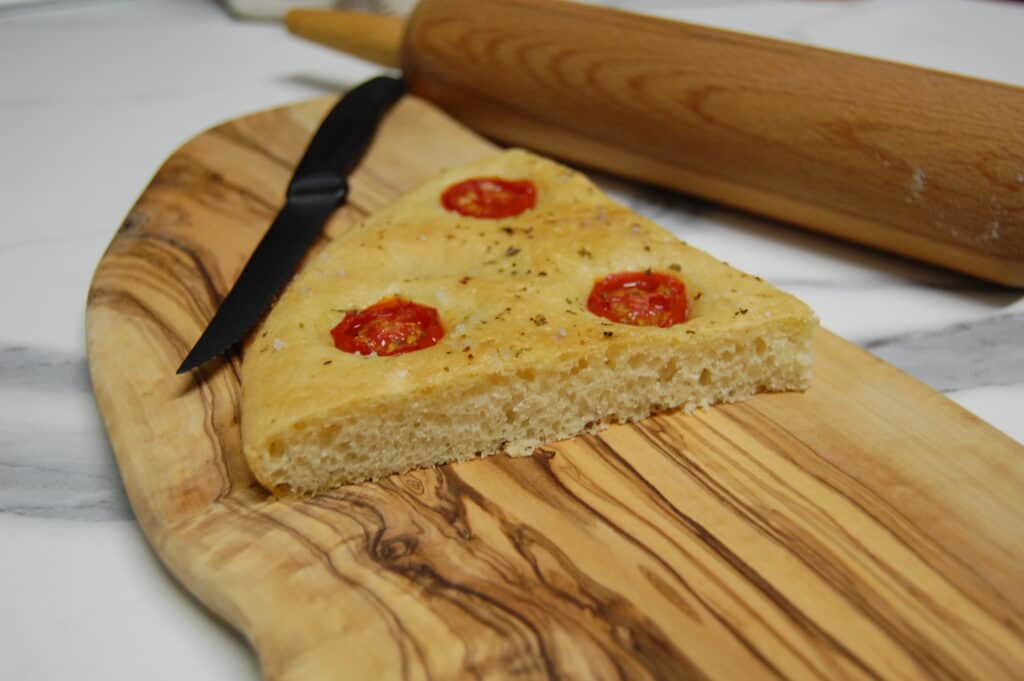 A slice of focaccia with cherry tomatoes and oregano and a knife on a wooden chopping board with a rolling pin in the background