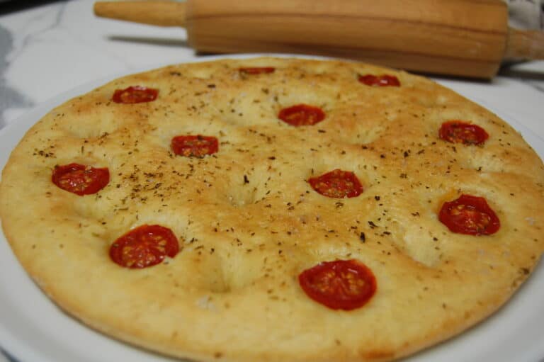 A close up view of a whole round cherry tomato and oregano focaccia with a rolling pin in the background