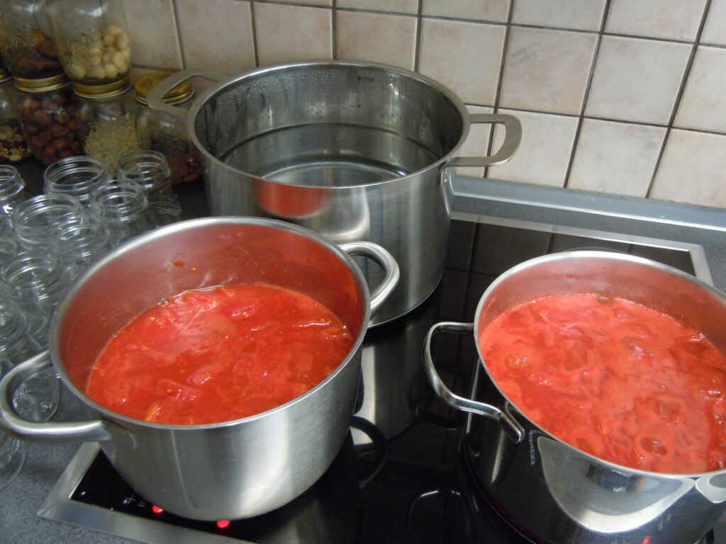 Two saucepans of tomato sauce boiling on the stove with a pan for sterilising them in the background