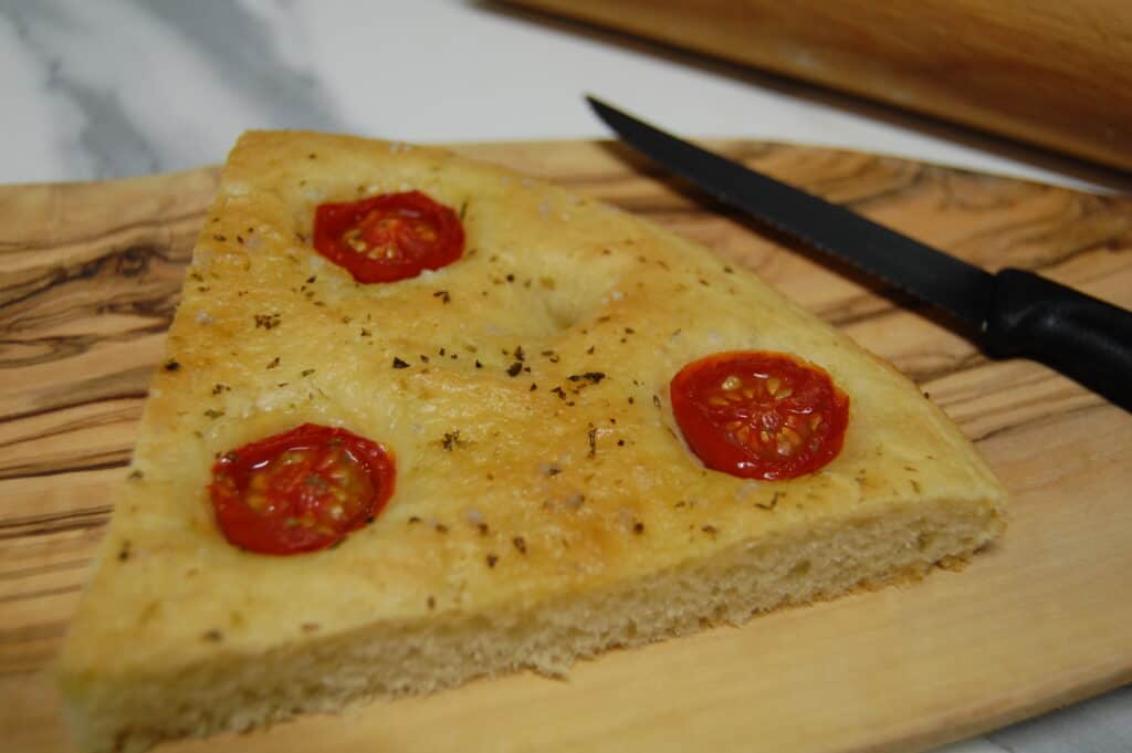 A close up image of a triangular slice of cherry tomato and oregano focaccia and a knife on an olive wood board