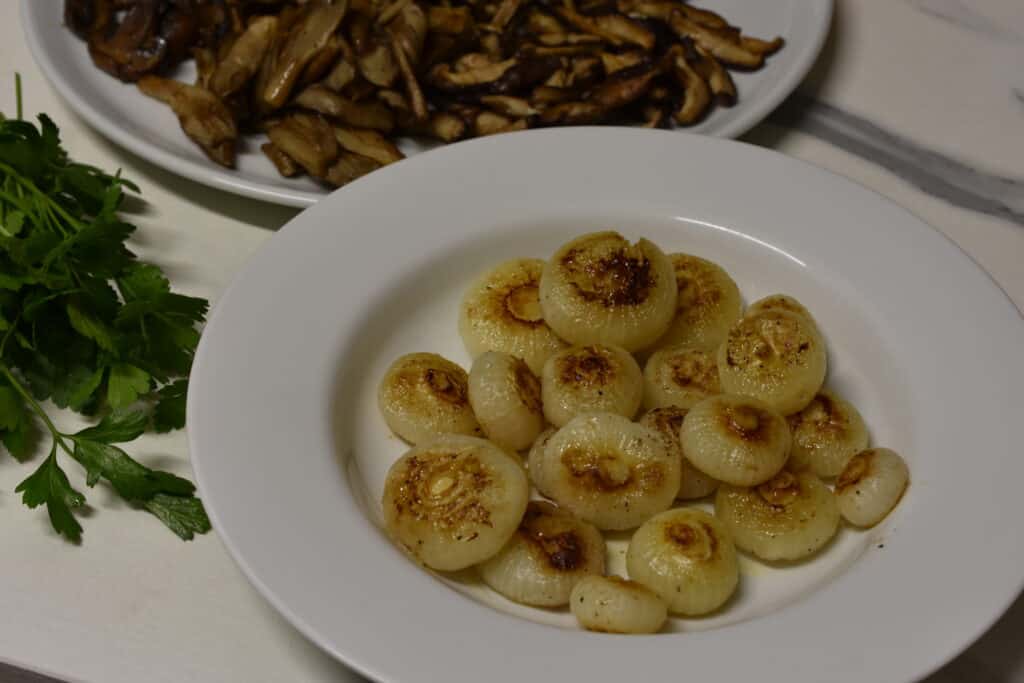 Fried borettane onions on a white plate on a kitchen worktop with some parsley and a plate of fried mushrooms in the background