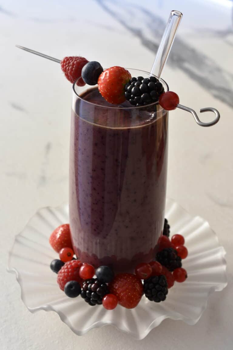 Mixed berry superfood smoothie in a glass with a skewer with berries on it and berries on a plate underneath