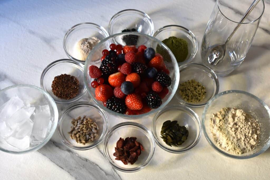 Strawberries, blueberries, raspberries, blackberries and redcurrants in a bowl, surrounded by smaller bowls containing superfoods, ice cubes, protein powder and a glass with a spoon on a worktop