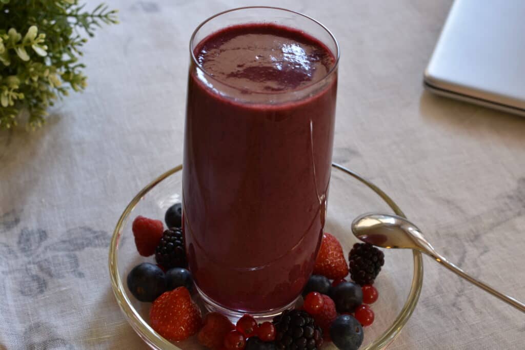 Mixed berry superfood smoothie on a glass plate with whole berries and a long spoon on a table cloth with props.