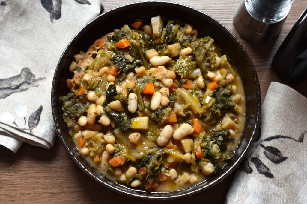 Ribollita toscana (tuscan ribollita bean soup) in a brown bowl on a table with kitchen cloths, a bottle of oil and a pepper mill