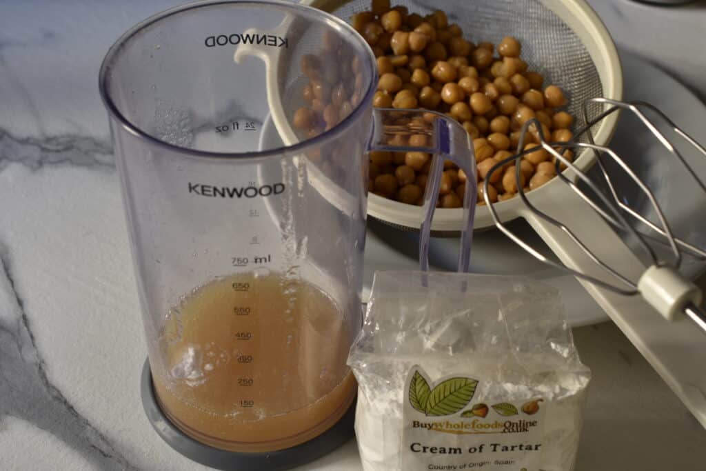 Aquafaba in a transparent plastic container with a packet of cream of tartar and chickpeas in a sieve in the background