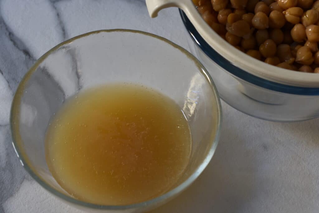 Aquafaba (the cooking liquid from chickpeas) in a glass bowl with a sieve full of chickpeas in the background