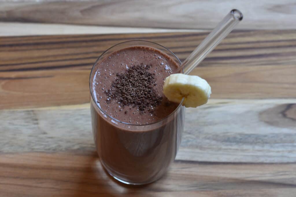 A glass of chocolate banana protein shake with grated dark chocolate, a slice of banana and a glass straw on a wooden board