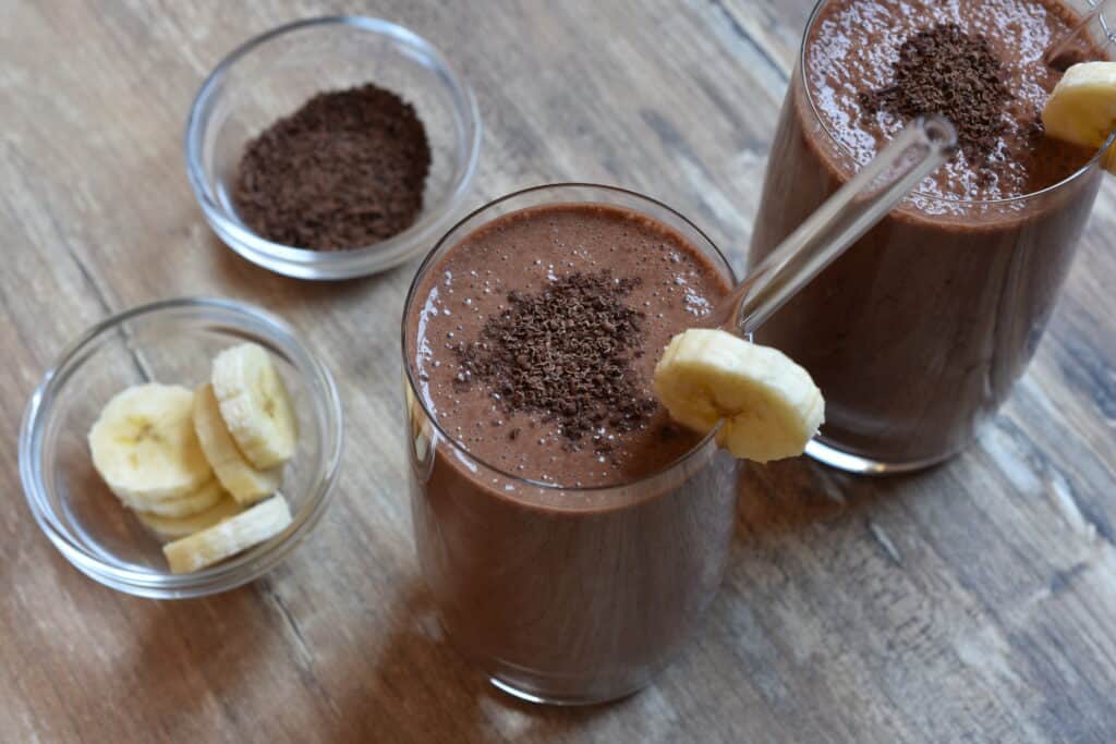 Close-up image of two glasses of chocolate and banana protein shake with garnish on a wooden table.