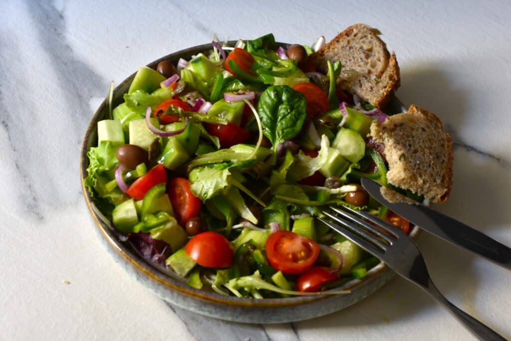 A partially-eaten Greek-style mixed salad on a grey plate with slices of bread and a knife and fork.