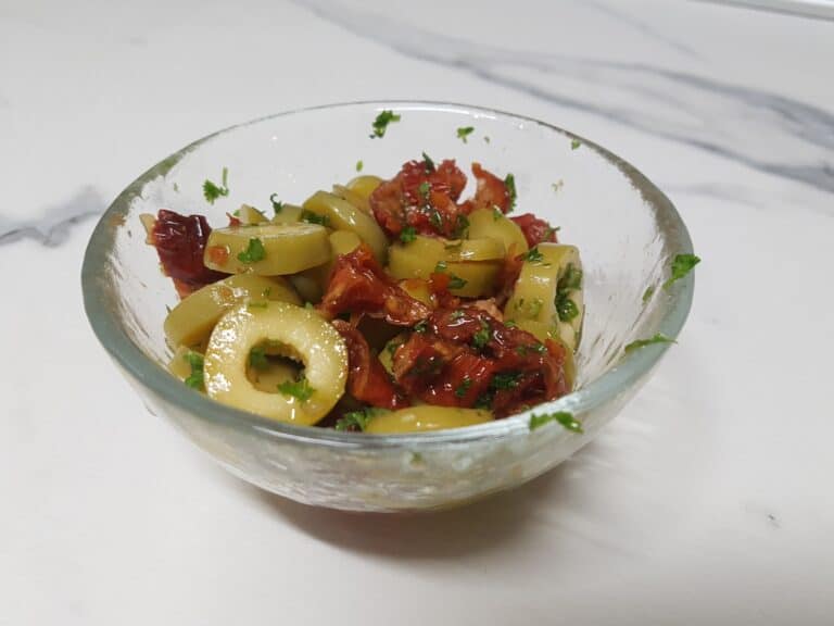 Green olive and sundried tomato garnish for roasted vegetable couscous in a small glass bowl on a kitchen worktop