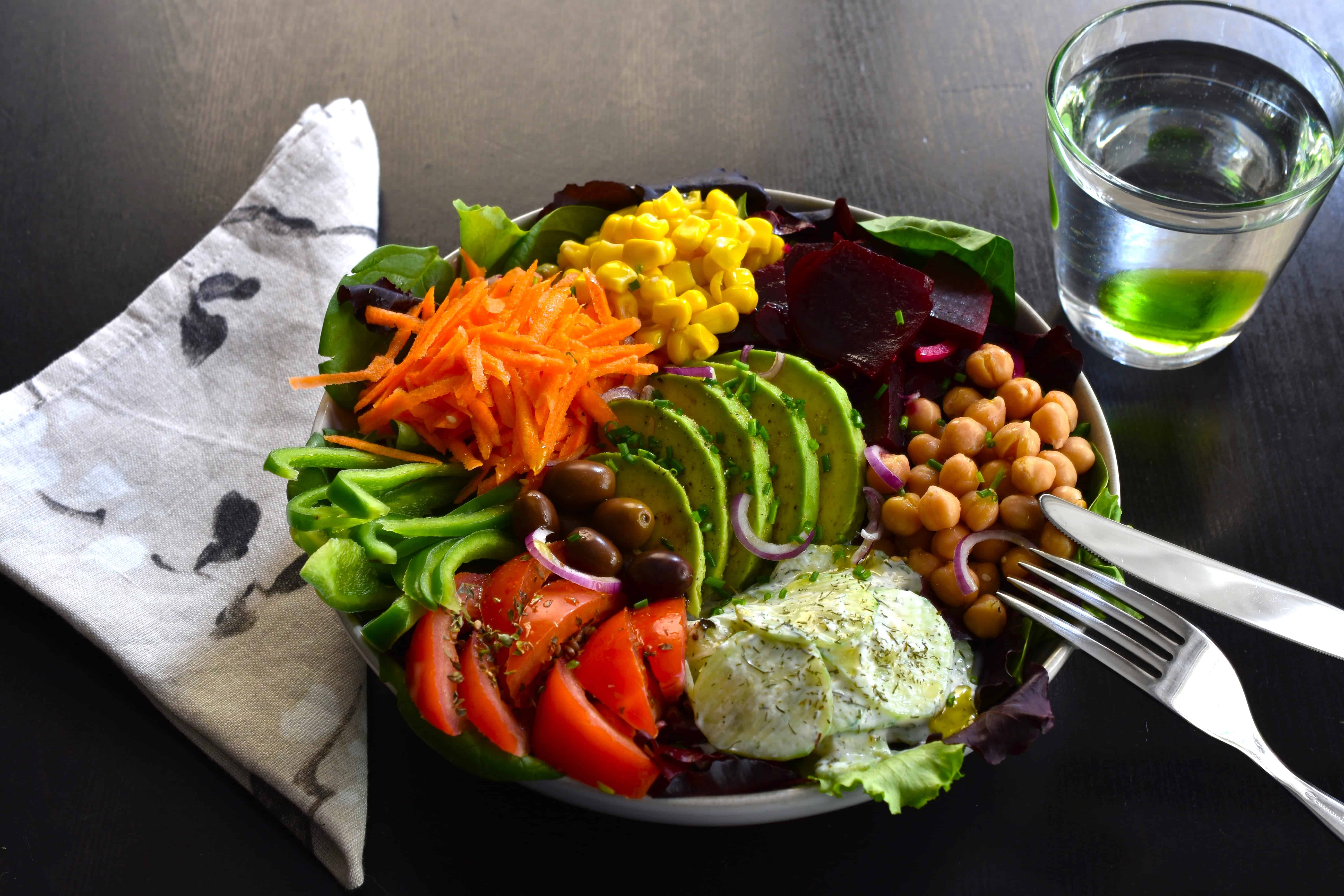 Rainbow mixed salad bowl with a knife and fork on a dark table with a napkin and a glass of water 