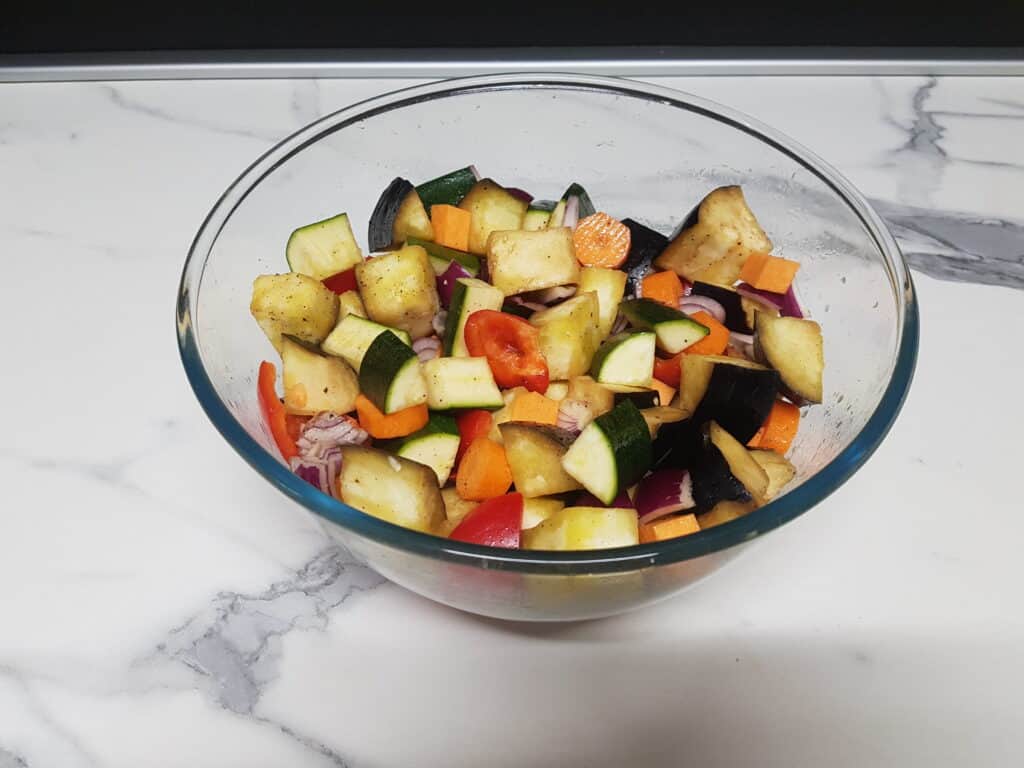 Cubes of raw vegetables tossed in olive oil and seasoned with salt and pepper in a glass bowl.