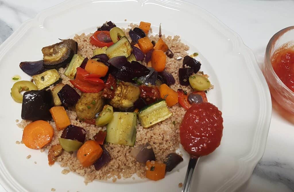 Roasted vegetable couscous on a white plate with a spoonful of sweet and sour tomato sauce hovering over the plate.