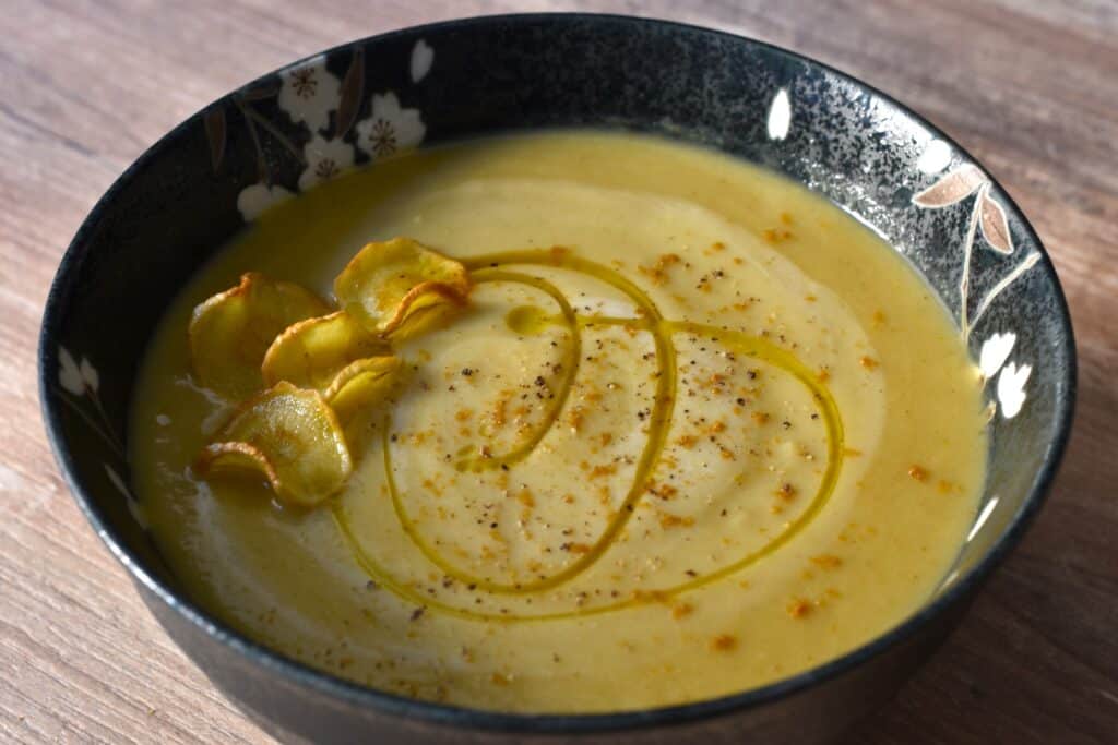 Spicy parsnip soup with a garnish of parsnip chips, ground cumin seeds and evo oil, in a decorative bowl on a wooden table.