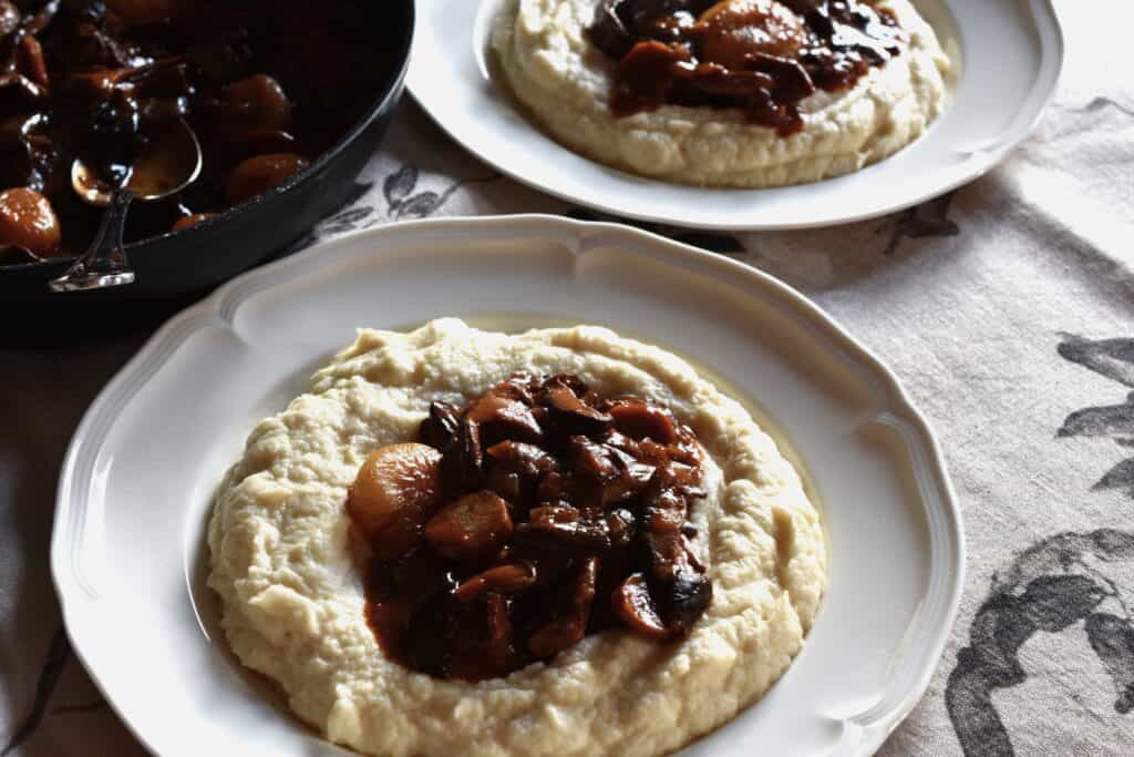 Two plates of mushroom bourguignon in wells of vegan celeriac mash on a tablecloth with a pan in the background,