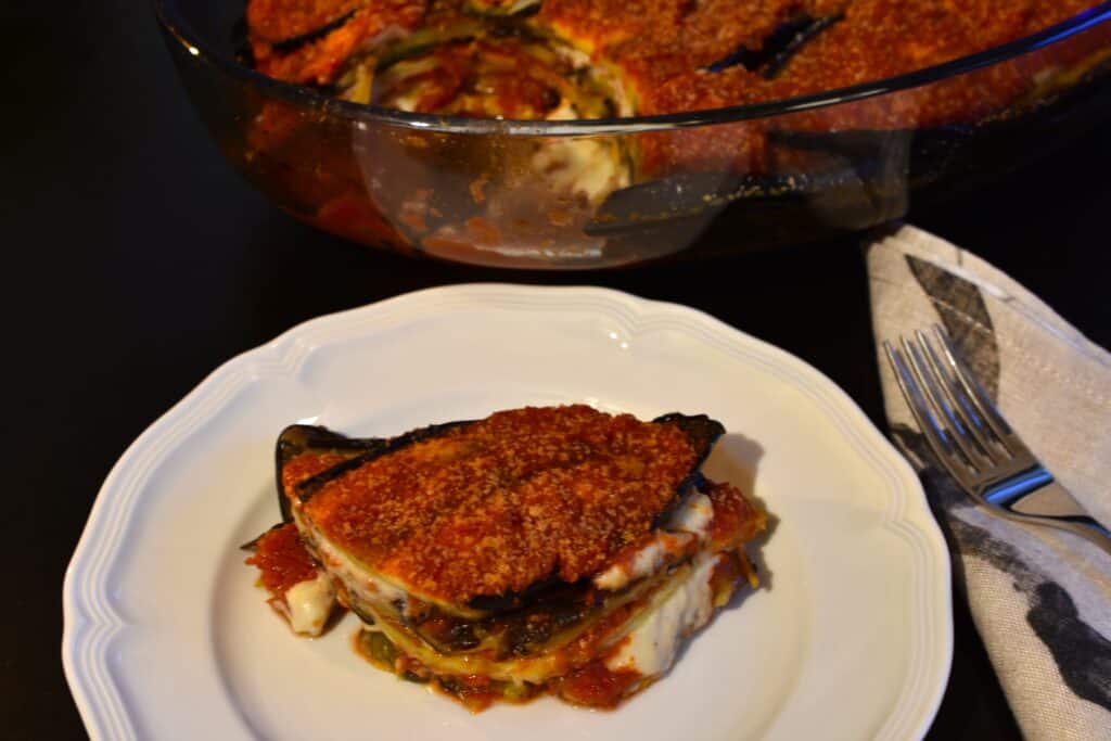 A portion of vegan aubergine parmigiana on a white plate with a serviette and fork and a dish of parmigiana in the background