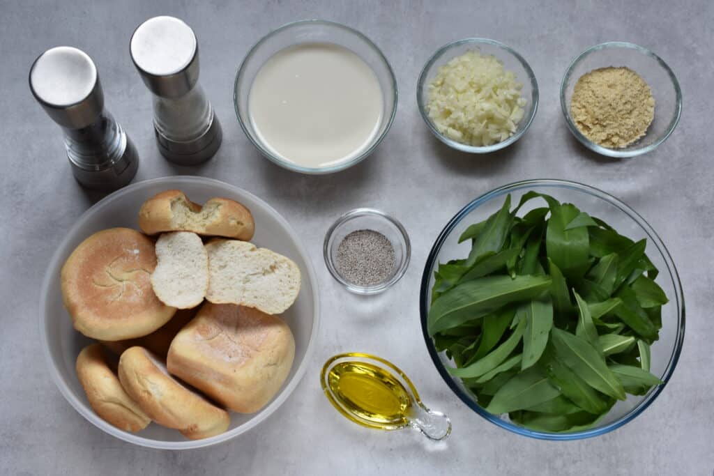 Ingredients for wild garlic bread dumpling: stale bread, soya milk, oil, onions, wild garlic, nutritional yeast, chia seeds, salt and pepper, in individual bowls.