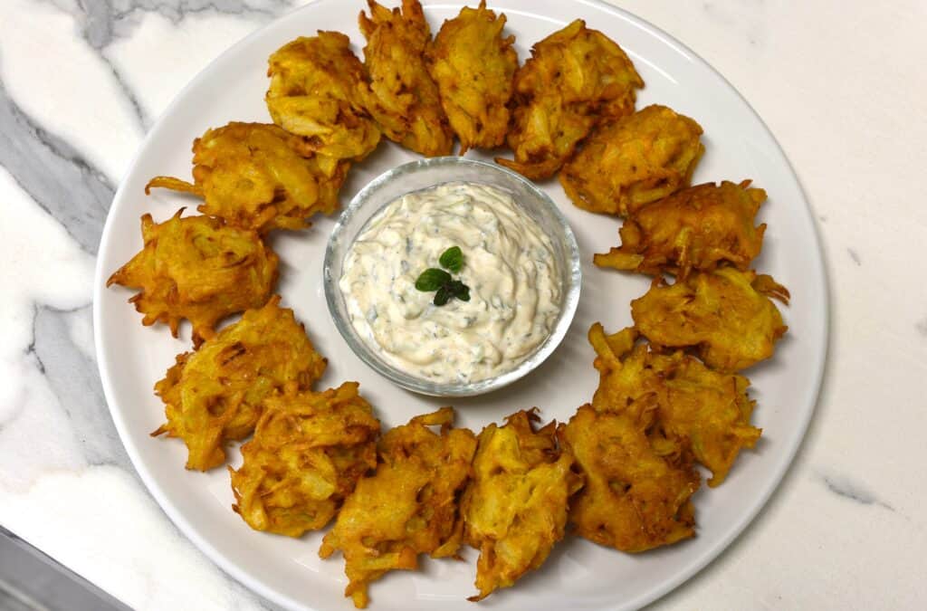 A ring of onion bhaji and a bowl of cucumber mint raita on a white plate.