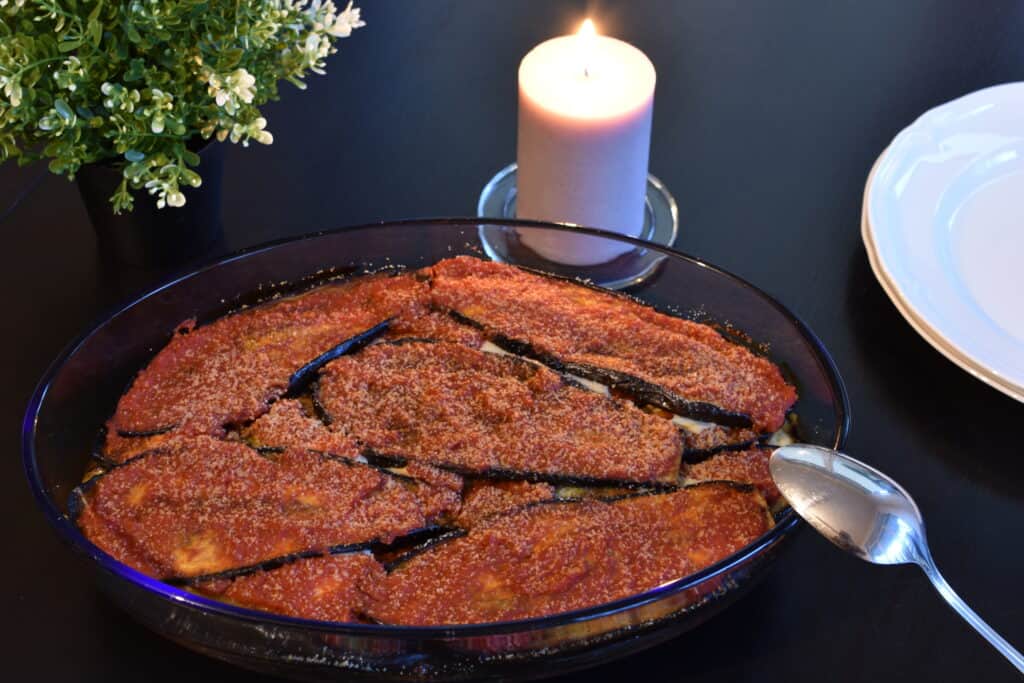 Vegan aubergine parmigiana in an oval glass oven dish with a spoon on a dinner table.