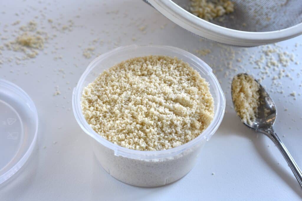 Vegan parmesan cheese in a plastic container with a spoon and a sieve on a kitchen worktop.