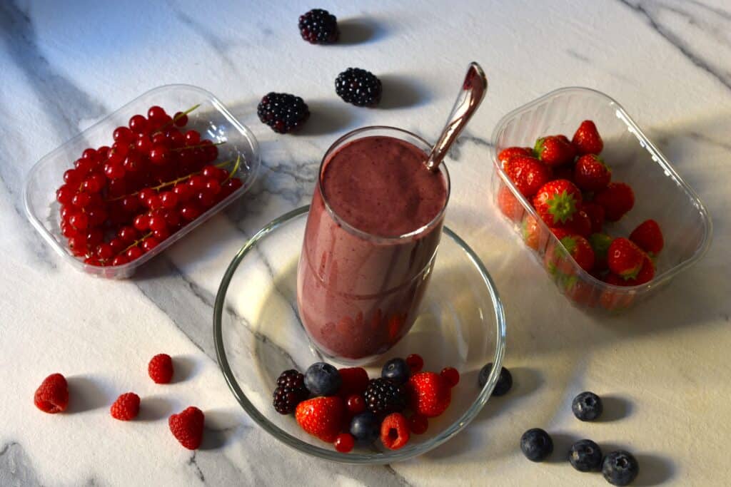 Mixed berry superfood smoothie in a glass with a long spoon and whole berries on a kitchen worktop.