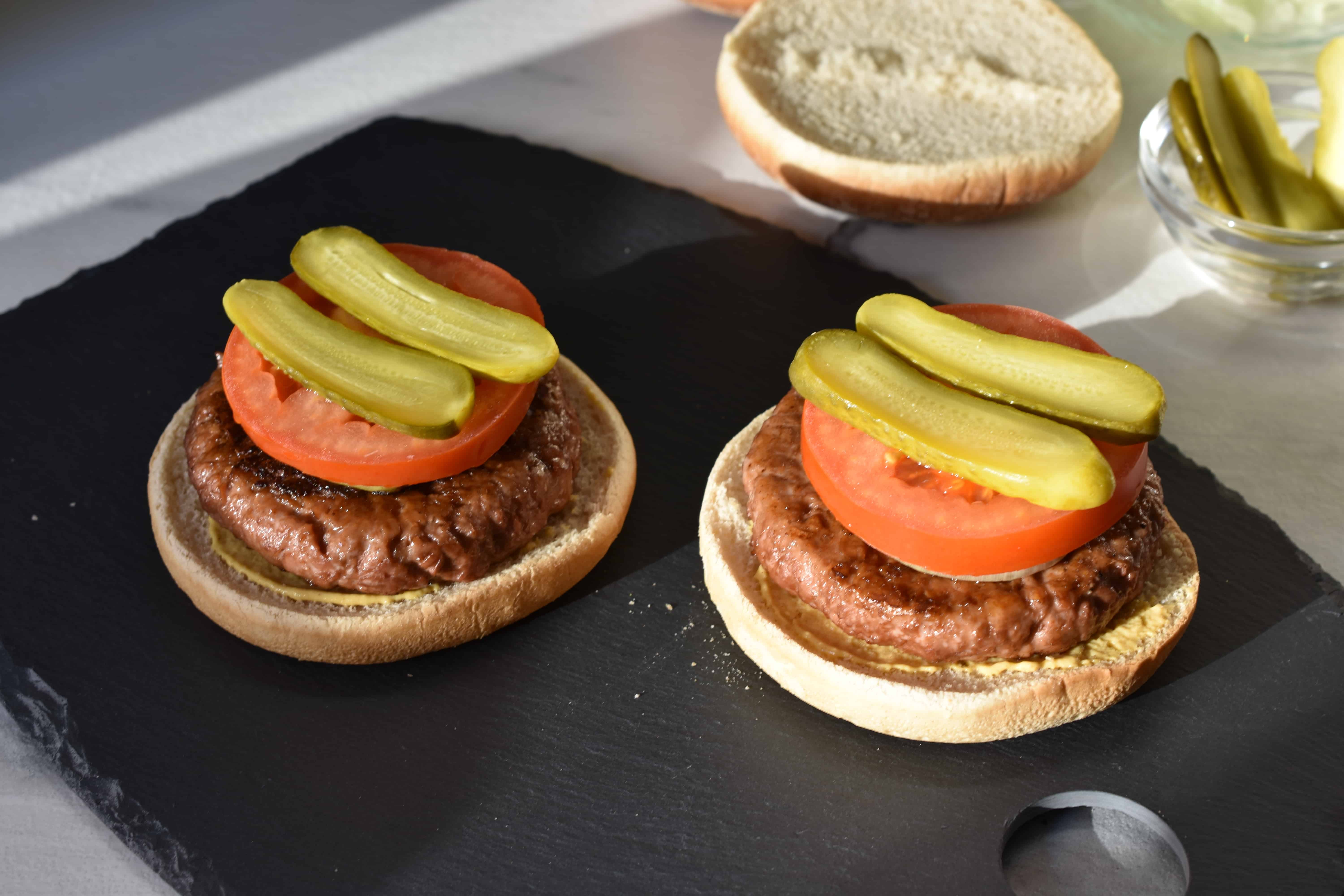 Vegan burger bun bottoms with a vegan fast food burger, fried onions, a slice of tomato and pieces of gherkin.