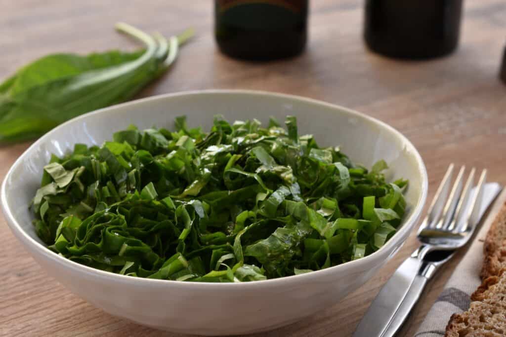 A close-up image of Italian spadona (sword) chicory in a white bowl with a knife and fork and wholemeal sourdough bread on a wooden table.