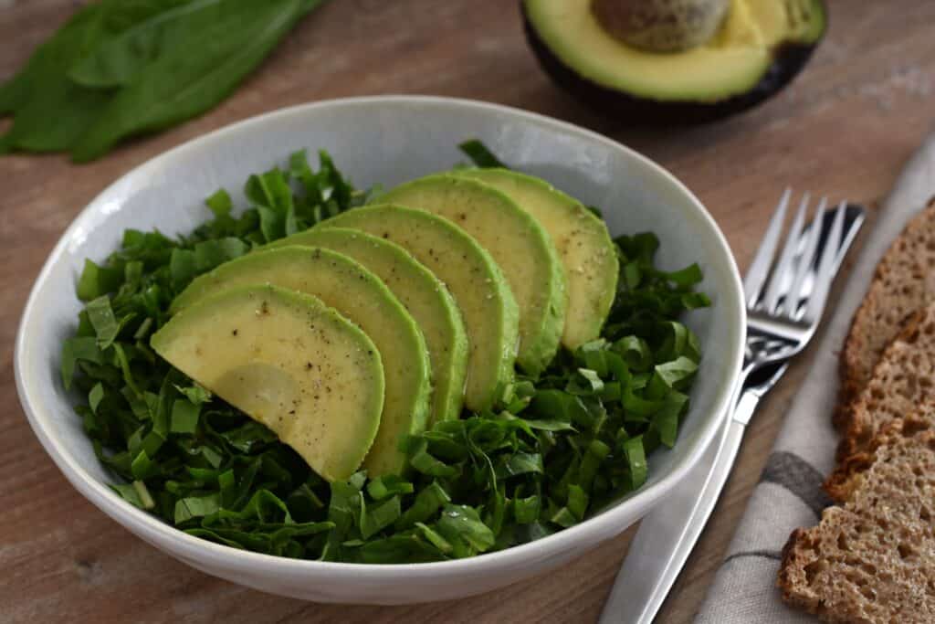 Italian spadona (sword) chicory and avocado salad in a white bowl with a knife and fork and wholemeal sourdough bread on a wooden table.