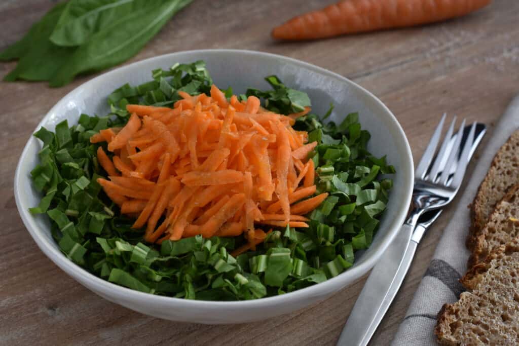 Italian spadona (sword) chicory and carrot salad in a white bowl with a knife and fork on a wooden table with slices of bread.