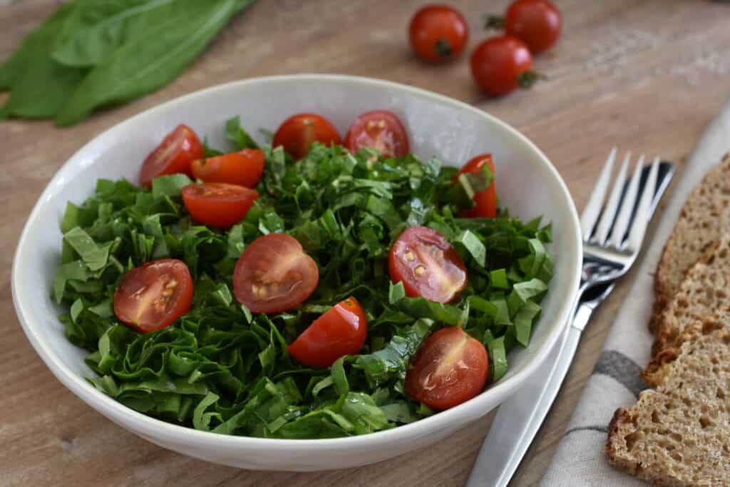 Italian spadona (sword) chicory and cherry tomato salad in a white bowl with a knife and fork and slices of sourdough wholemeal bread.