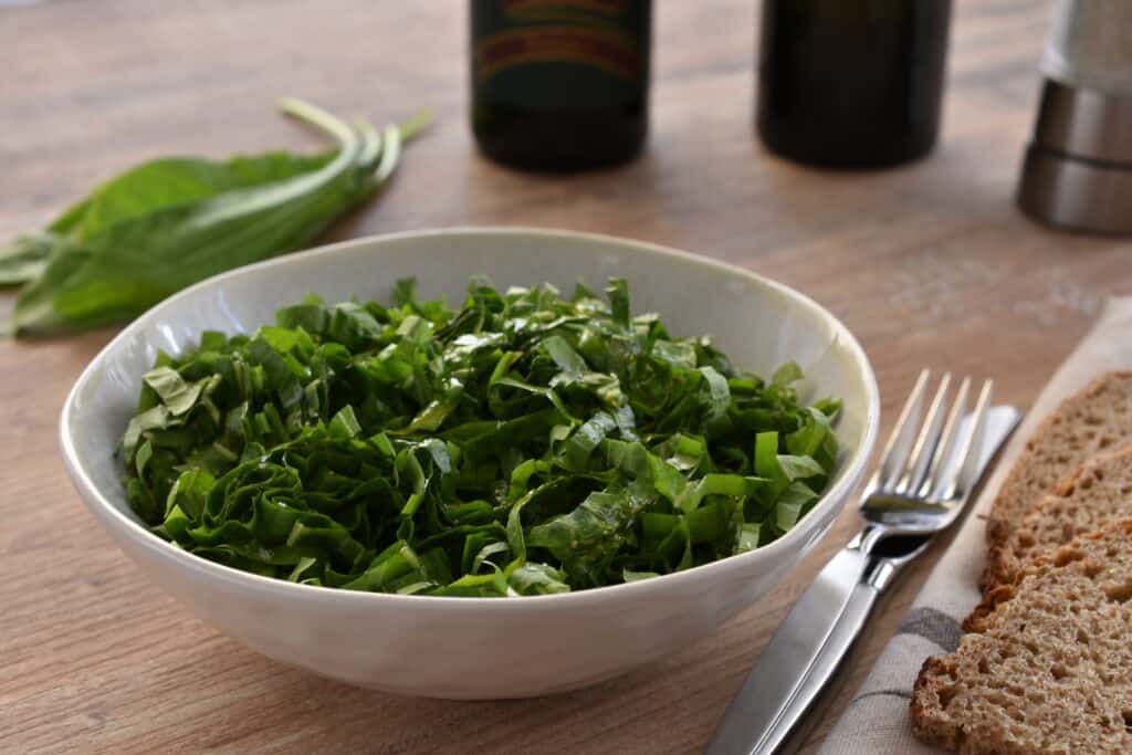 Italian spadona (sword) chicory salad in a white bowl with a knife and fork and wholemeal sourdough bread on a wooden table.