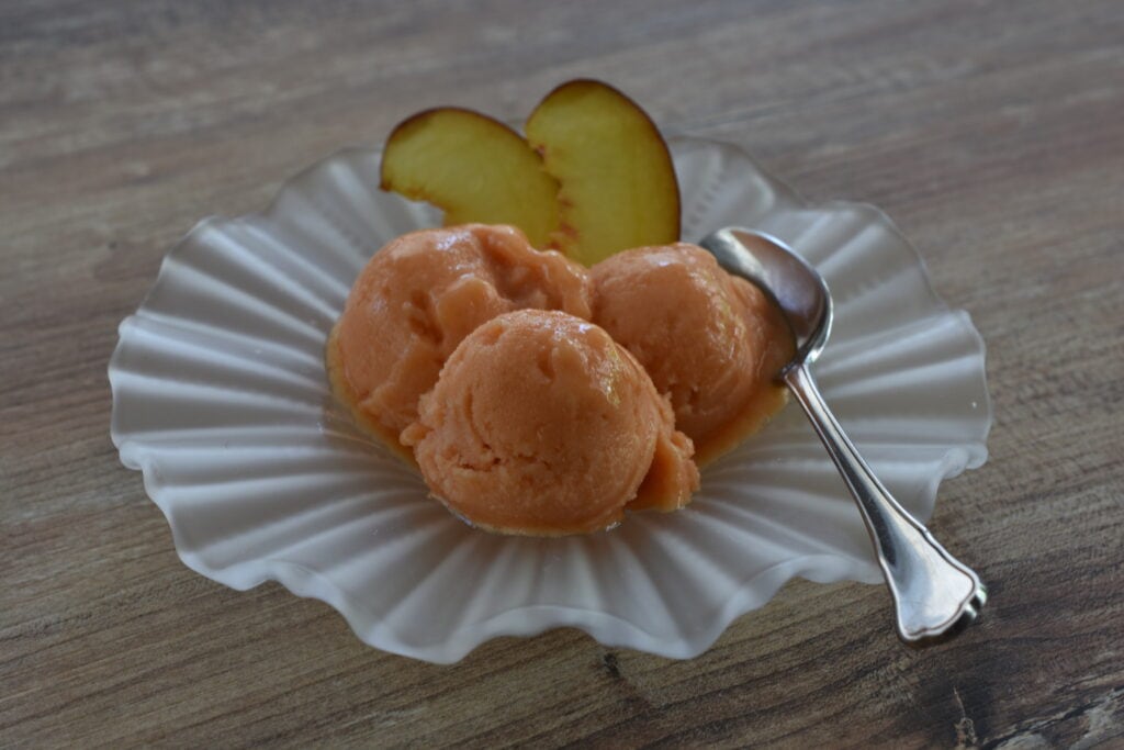 Three scoops of peach sorbet in a glass dish with a spoon and slices of peach.