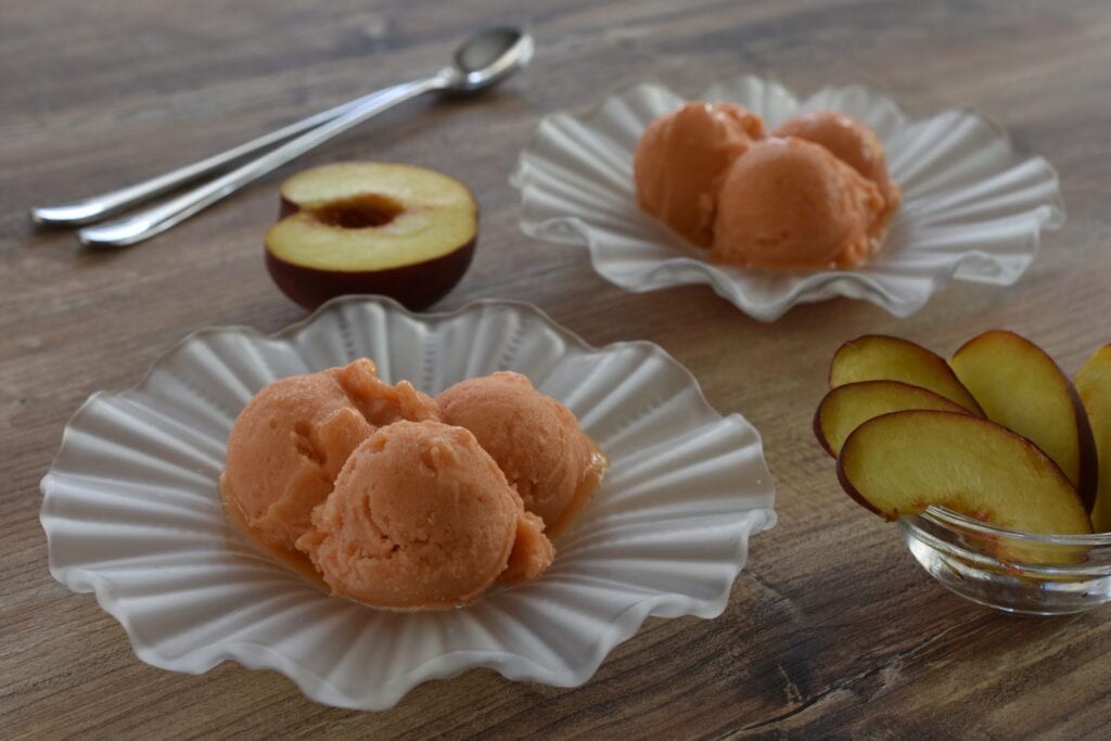 Scoops of peach sorbet in glass ice cream dishes on a wooden table, with spoons and slices of peach.