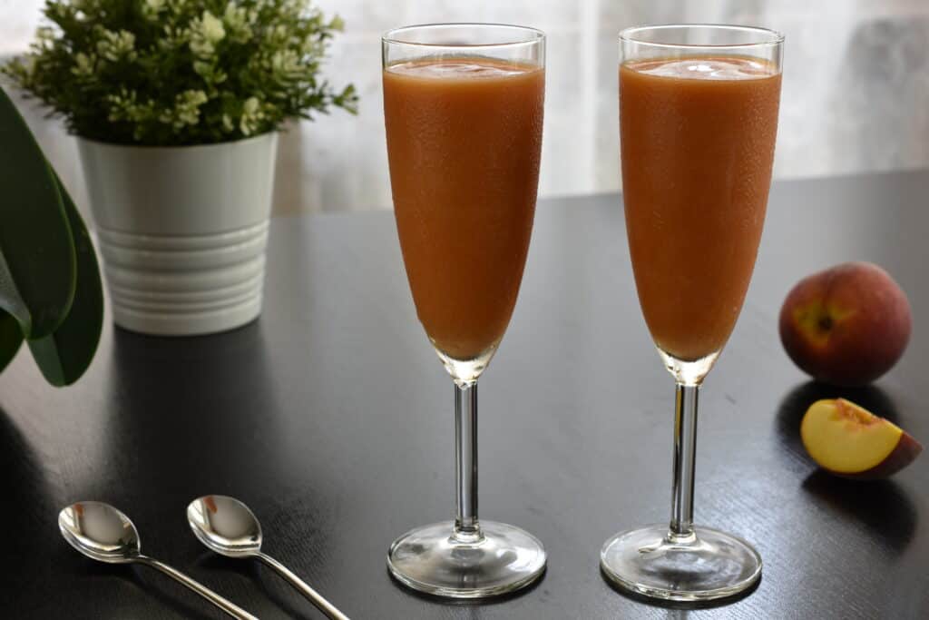 Two flute glasses of peach sorbet on a table with spoons and pieces of peach.