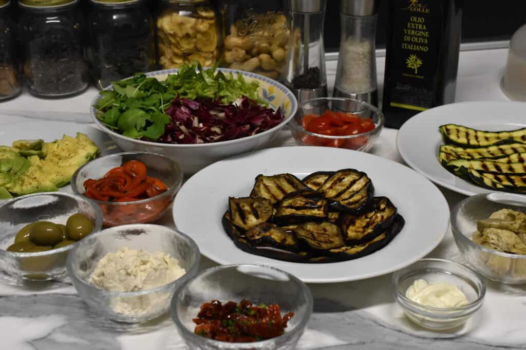 Ingredients for vegan piadinas: grilled courgettes, grilled aubergines, sautéed red peppers, avocado, lettuce, radicchio, rocket, grilled artichokes, sundried tomatoes, olives, cashew cream cheese, vegan mayonnaise, in bowls on a kitchen worktop.