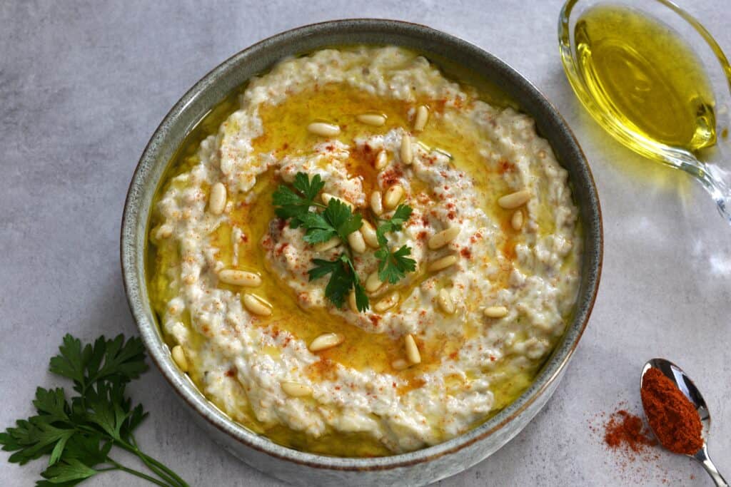 A bowl of vegan baba ganoush (moutabal) with a garnish of pine nuts, extra virgin olive oil, paprika and parsley  on a table with props.