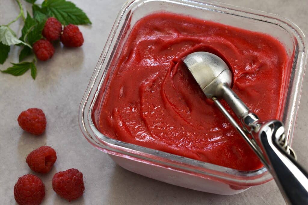 Raspberry sorbet in a glass container with a scoop.