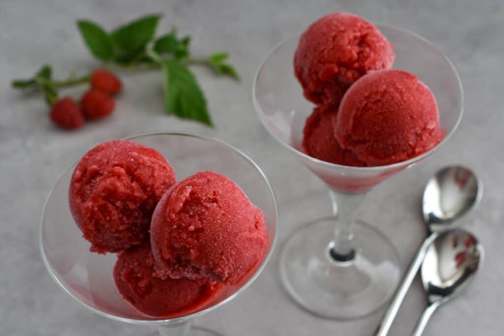 Scoops of raspberry sorbet in glass bowls on a table with spoons.