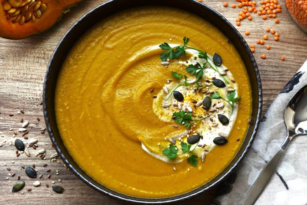 A bowl of pumpkin and red lentil soup with a garnish of coconut milk, mixed seeds, parsley and chili.