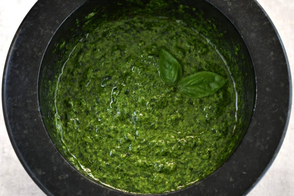 A close-up image of vegan basil pesto in a mortar with a pestle and a garnish of basil leaves.