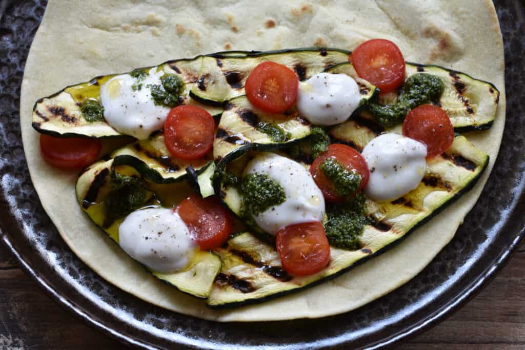 A piadina with grilled courgettes, cherry tomatoes, vegan stracchino, and basil pesto on a plate.