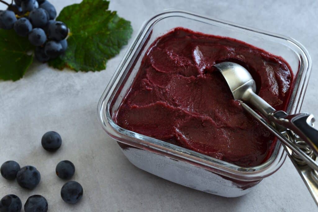 Concord grape sorbet in a glass container with an ice cream scoop.