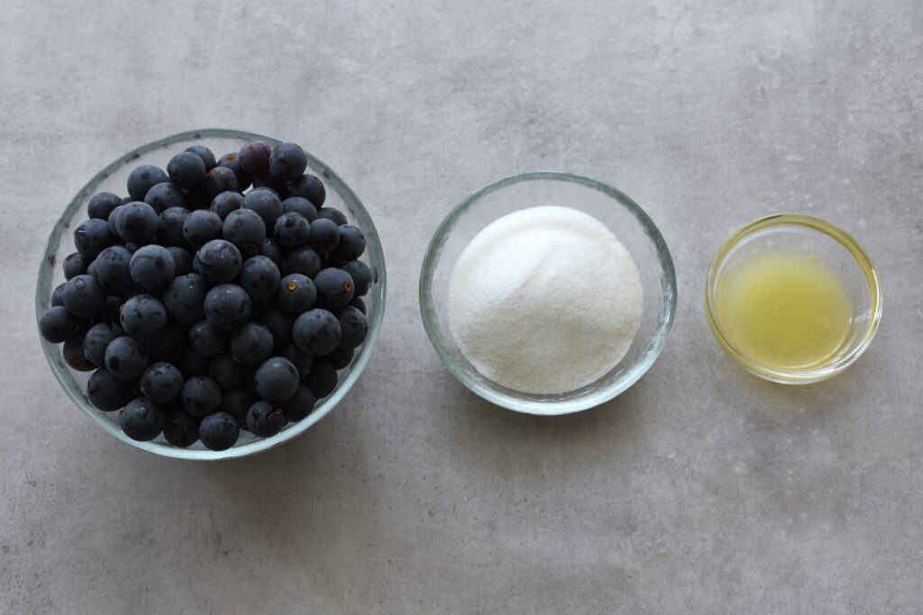 Concord grapes, sugar and lemon juice in glass containers on a table.