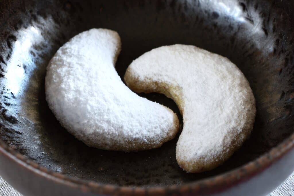 A close-up image of two vegan vanilla crescent cookies in a brown bowl.