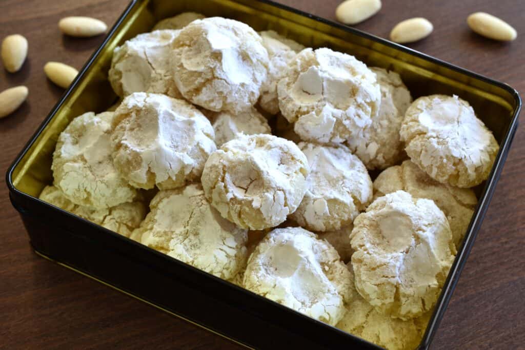 Vegan Italian almond cookies (pizzicotti alle mandorle) in a biscuit tin on a wooden table with almonds.