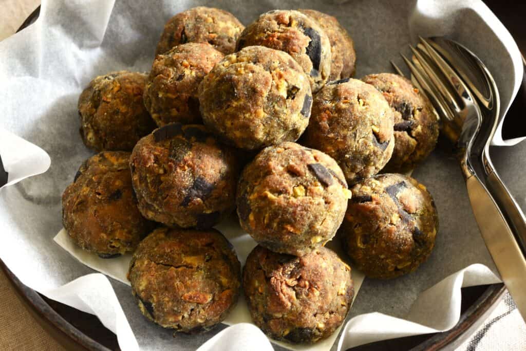 A close-up image of baked polpette di melanzane vegan eggplant meatballs in a serving dish with a spoon and fork.