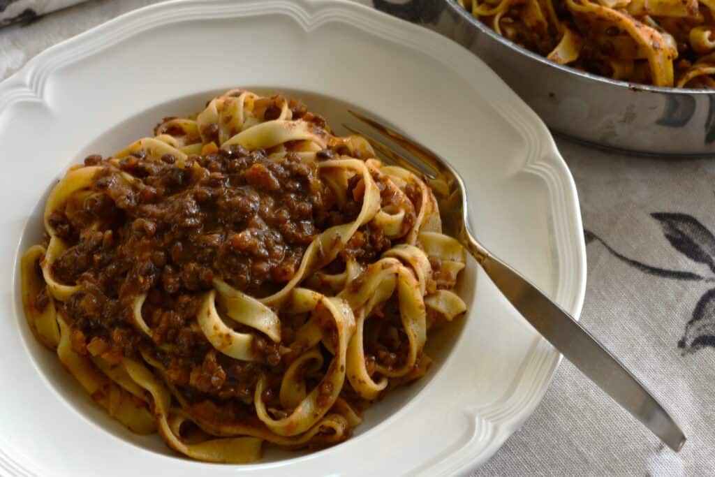 A close-up image of tagliatelle with vegan ragu on a white plate with a fork and a pan in the background.