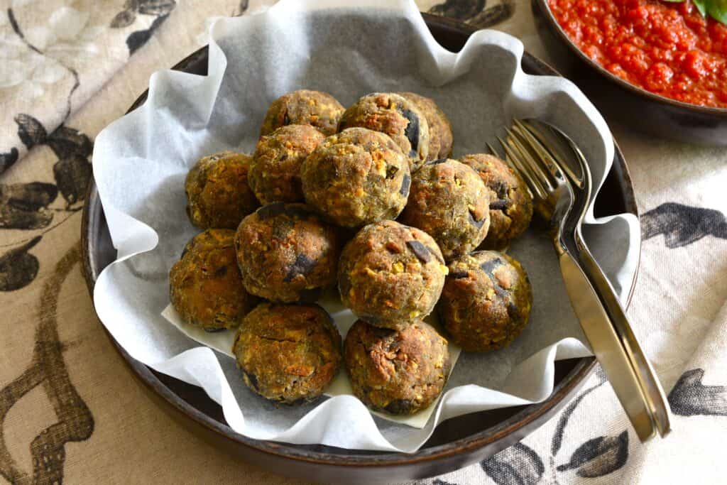 Polpette di melanzane vegan Italian eggplant balls in a bowl with a serving spoon and fork on a table with a bowl of tomato sauce