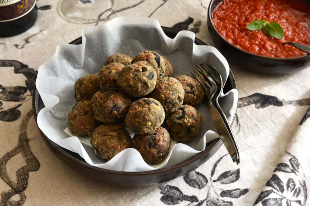 Polpette di melanzane vegan in a bowl with a serving spoon and fork on a table with tomato sauce and a bottle of wine.